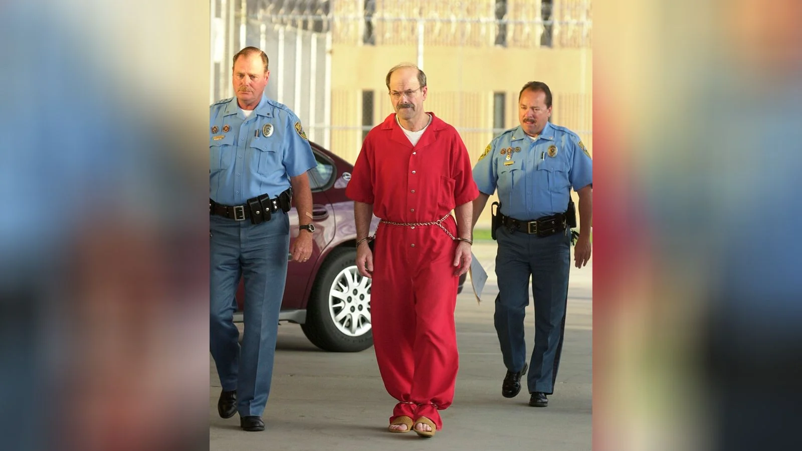 Dennis Rader, in a red jail jumpsuit, escorted by two officers