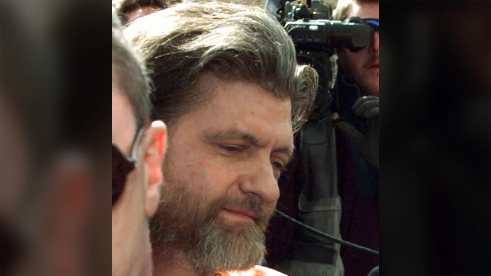 Ted Kaczynski, bearded and in an orange jail jumpsuit, escorted past news cameras