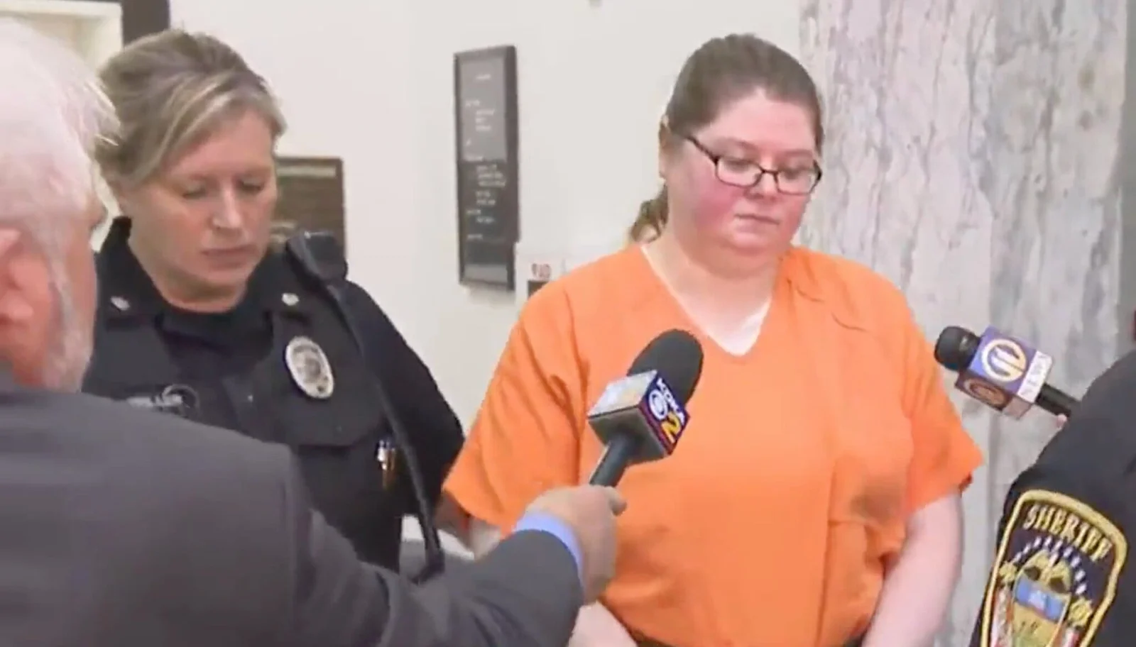 Heather Pressdee, in orange jail clothes and glasses, escorted by officers past reporters