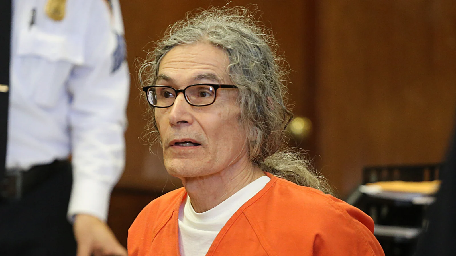 Rodney Alcala, gray-haired and in an orange jail uniform, seated at a defense table in court