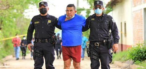 Hugo Osorio, in a blue shirt and red shorts, led by two masked Salvadoran police officers past crime-scene tape in Chalchuapa