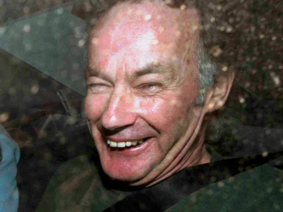 Photograph of Ivan Milat smiling through a car window, a corrections officer seated behind him