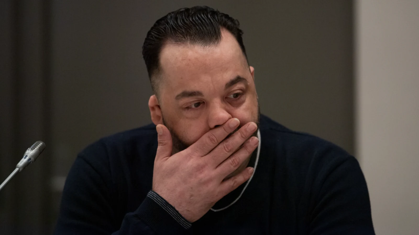 Niels Högel in a dark sweater, his hand over his mouth, seated beside a microphone in a courtroom