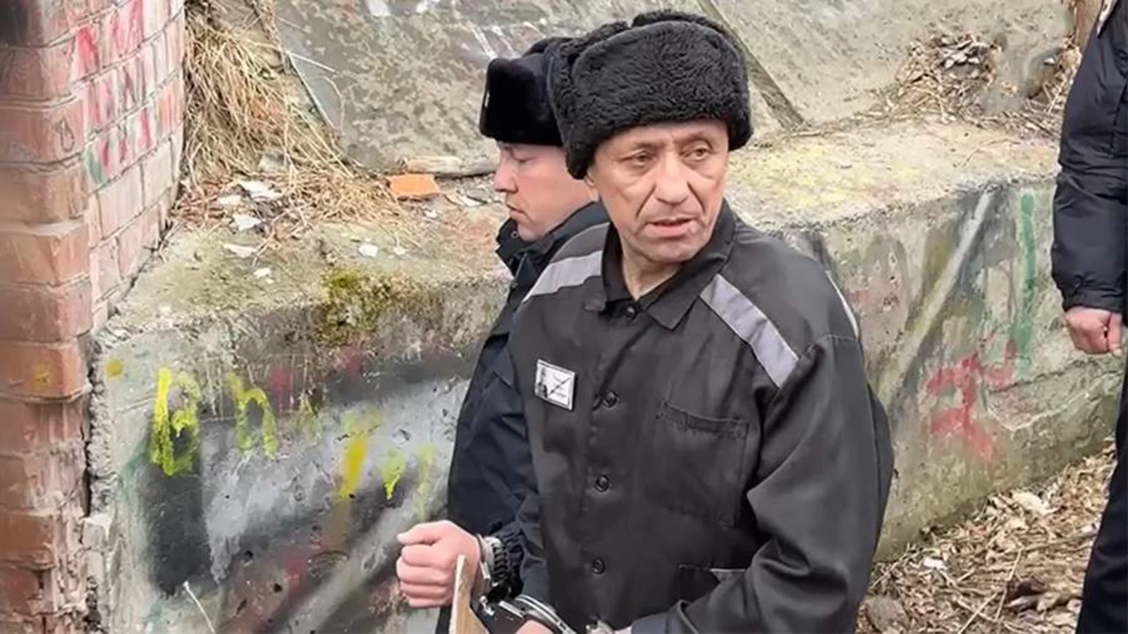 Mikhail Popkov, handcuffed in a black prison uniform and fur hat, under guard at an outdoor site