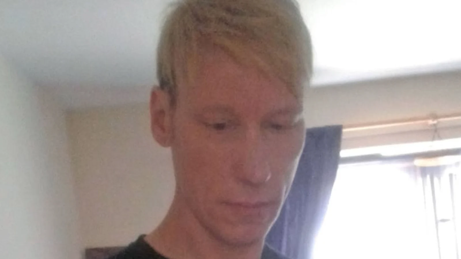 Stephen Port, blond-haired, in a black T-shirt, in a photograph taken indoors beside a curtained window