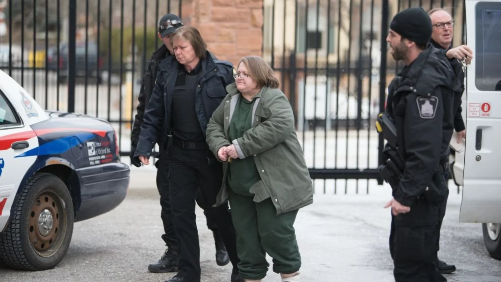 Elizabeth Wettlaufer in a green jacket, escorted by police officers past a police car outside a courthouse