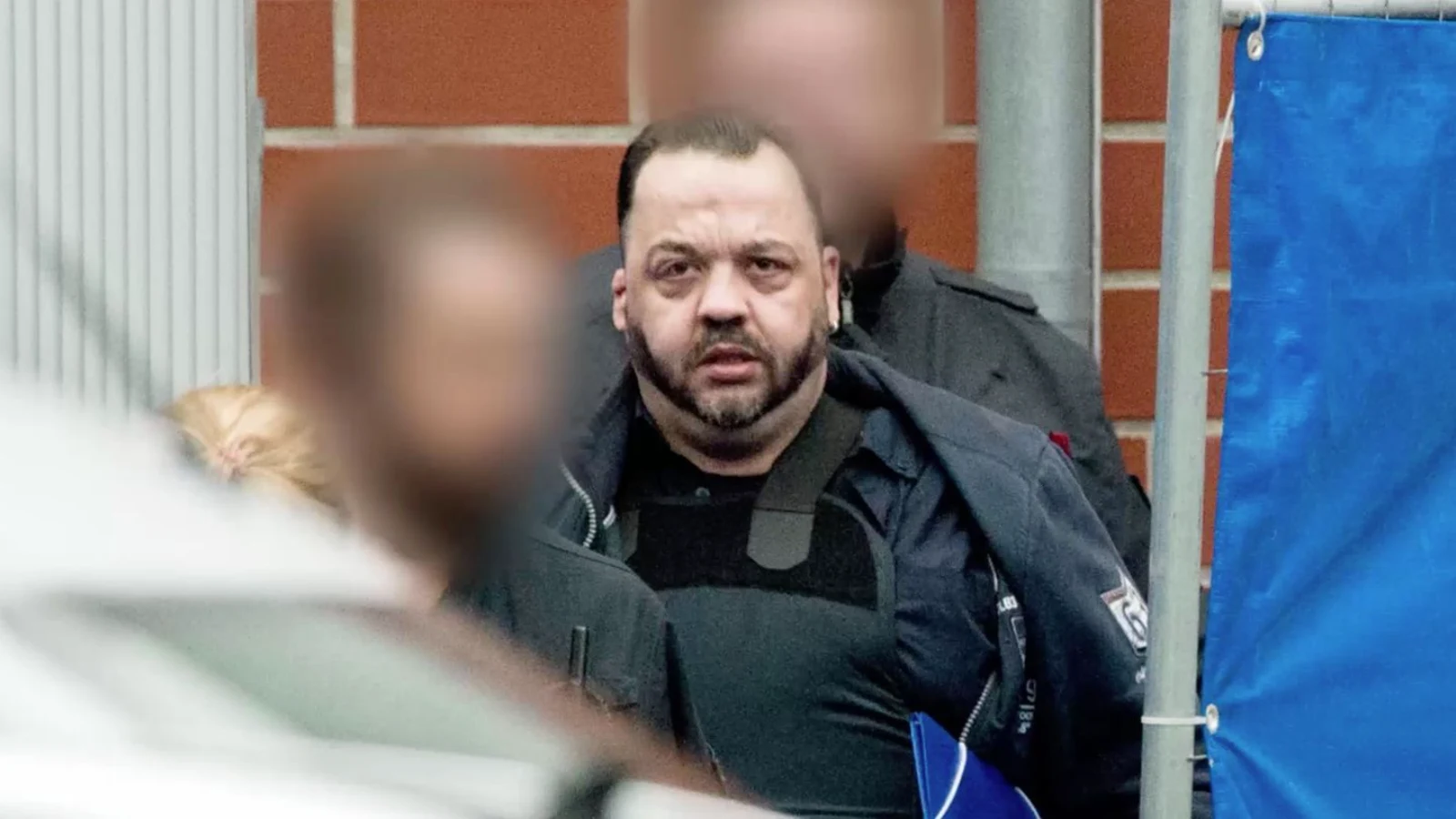 Niels Högel in a dark jacket, escorted by officers past a brick wall and a blue privacy screen