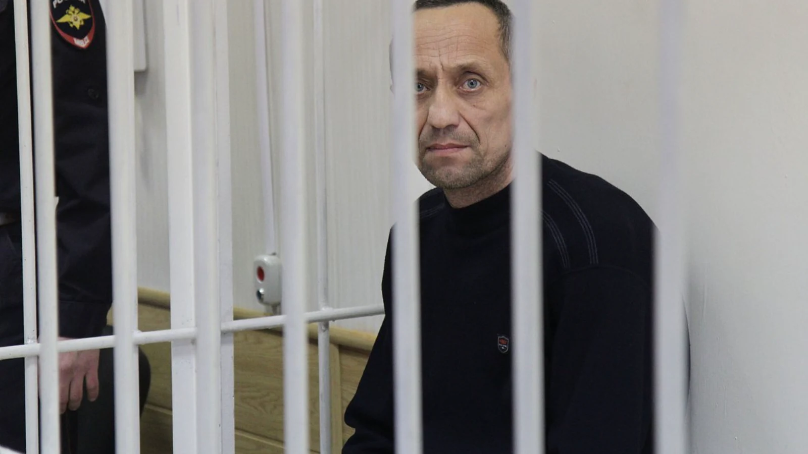 Mikhail Popkov in a dark sweater standing behind the white bars of a Russian courtroom cage, a uniformed guard at the edge of the frame