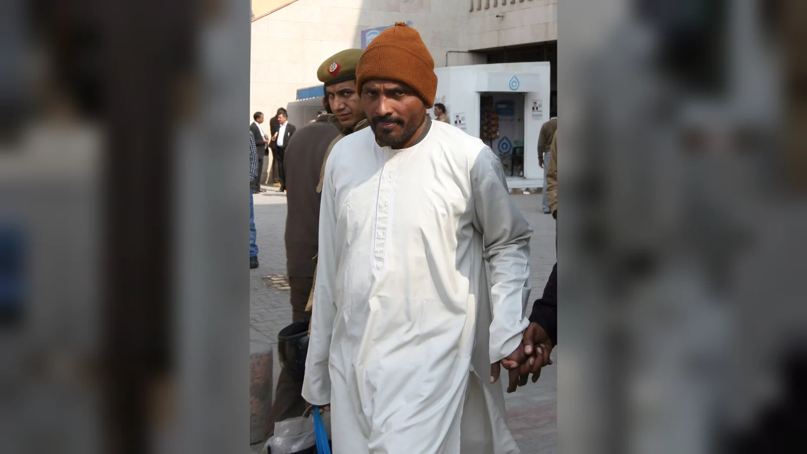 Chandrakant Jha, in a white kurta and a brown knit cap, escorted by a Delhi police officer