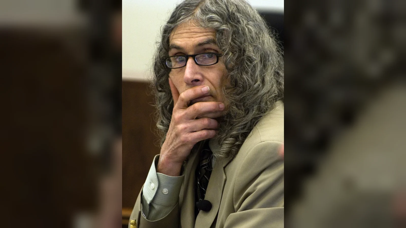Rodney Alcala, gray-haired and in glasses, in a courtroom with his hand to his face