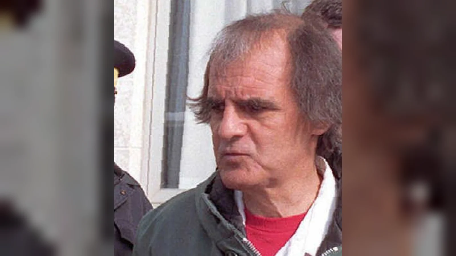 An older, gray-haired Clifford Olson in a dark jacket, escorted outside a building