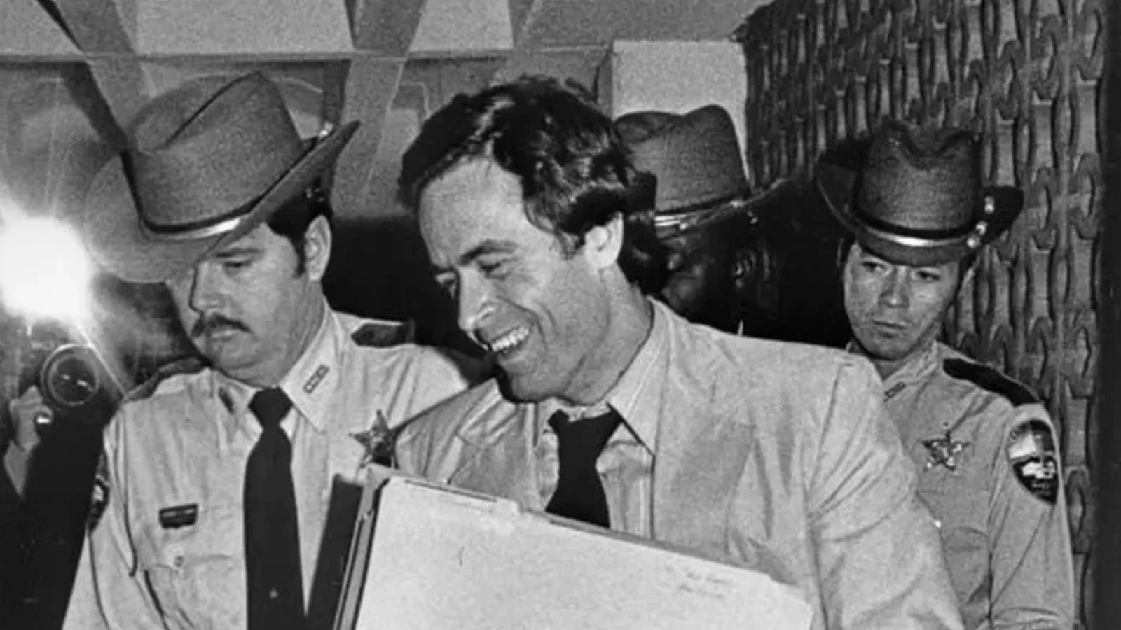 Ted Bundy smiling and carrying a box of papers as sheriff's deputies escort him