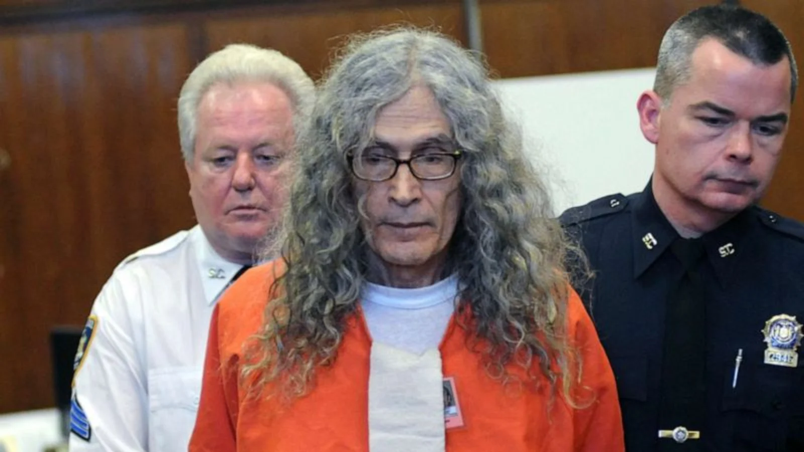 Rodney Alcala, with long gray hair and glasses, in an orange jail jumpsuit between court officers in Manhattan