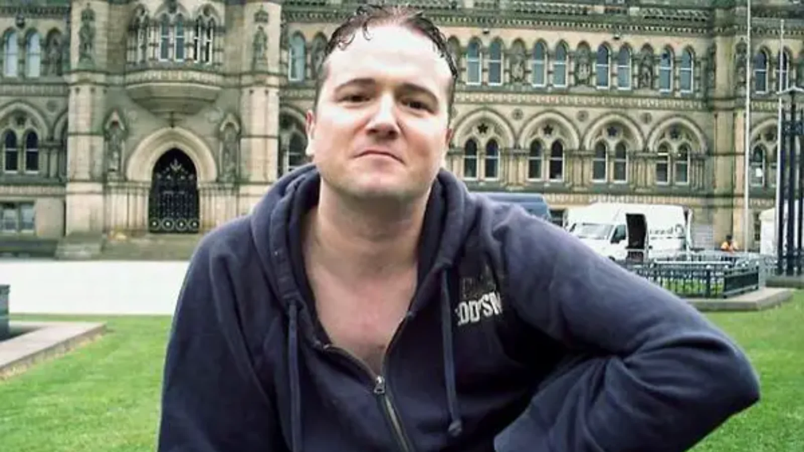 Stephen Griffiths in a dark hooded top, crouching on a lawn in front of Bradford City Hall