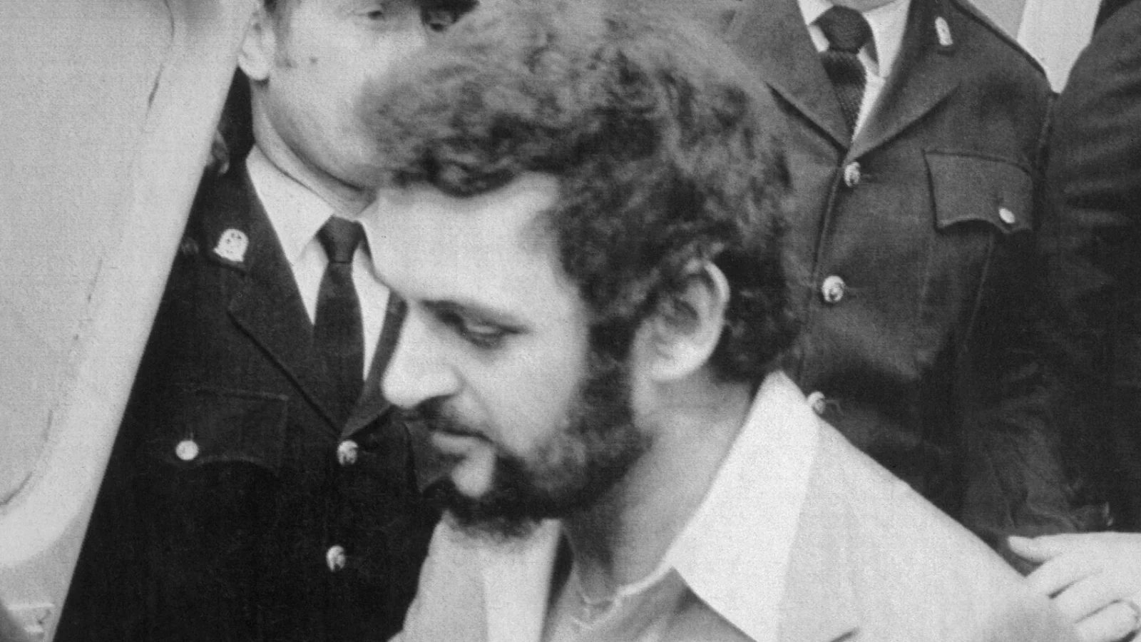 Peter Sutcliffe, bearded and looking down, led by police officers from a magistrates' court in 1981