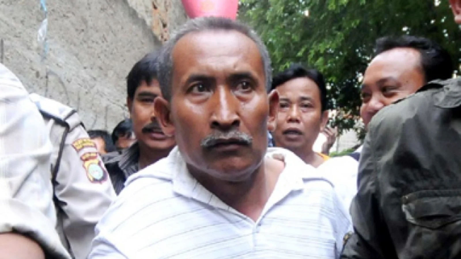 Baekuni in a white striped shirt escorted by Indonesian police through a crowd