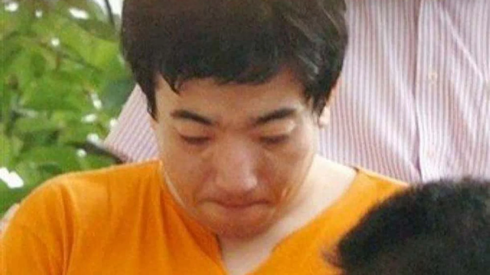 Hiroshi Maeue in an orange T-shirt, head lowered, escorted by police in Osaka