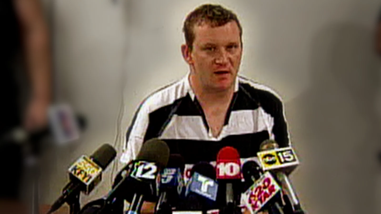 Dale Hausner in a striped jail uniform speaking at a jailhouse news conference behind television microphones