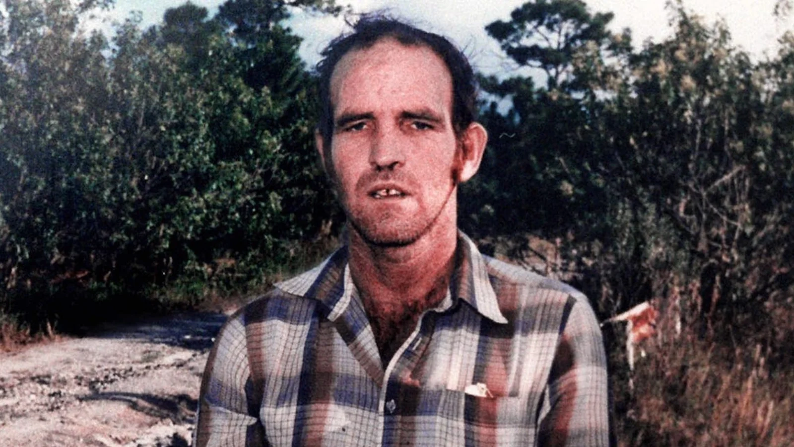 Ottis Toole in a plaid shirt standing beside a dirt road in Florida scrubland