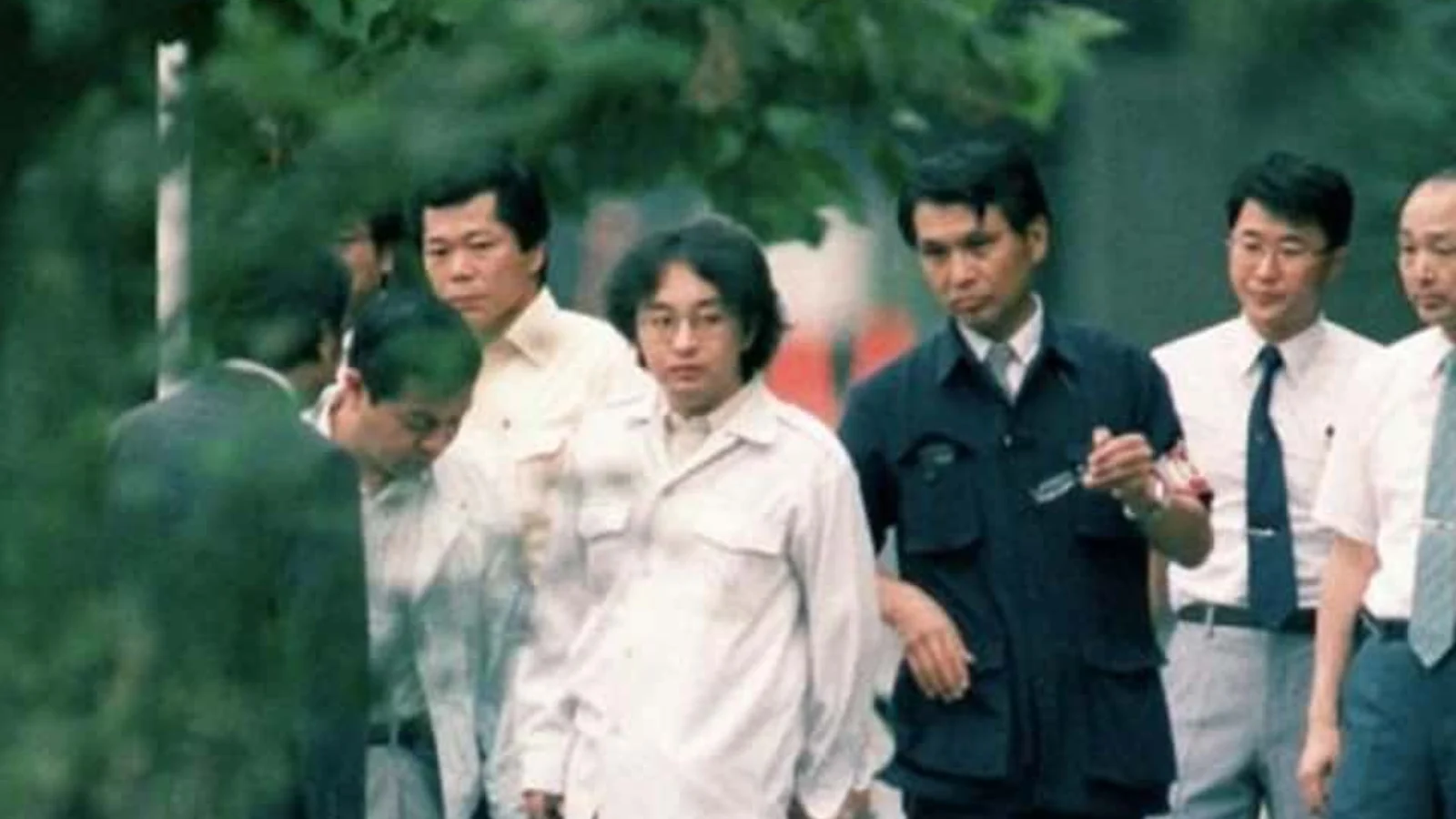 Tsutomu Miyazaki in a white shirt walking with investigators along a tree-lined path in 1989