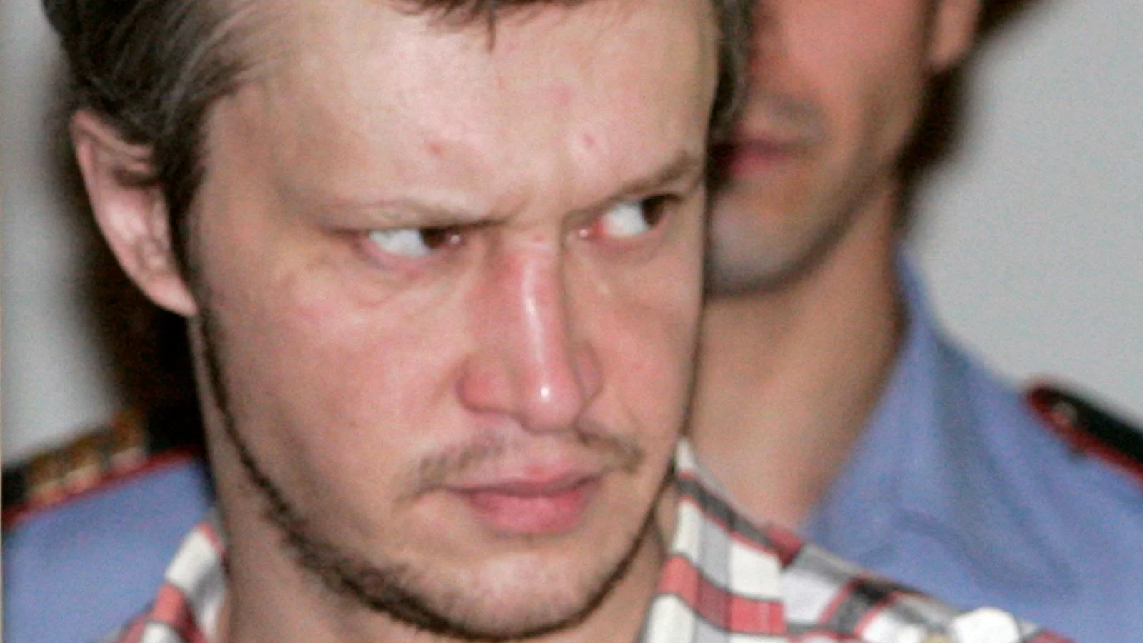 Alexander Pichushkin in a checked shirt glancing sideways, a police officer behind him, at a Moscow court