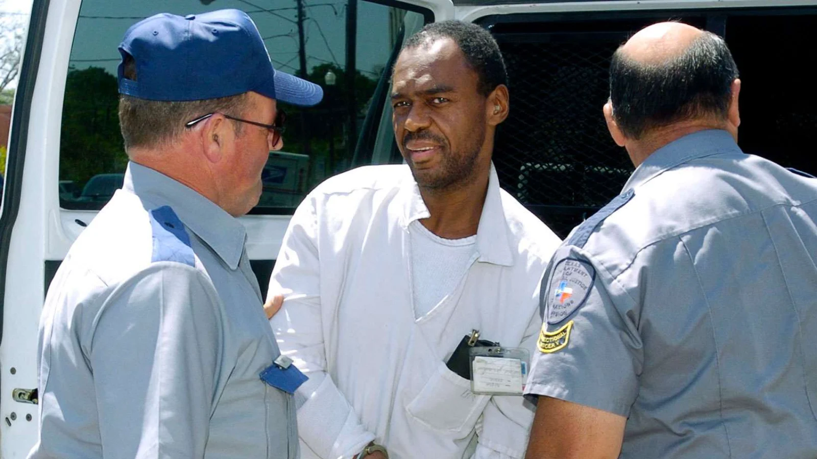 Coral Eugene Watts in white prison clothes, handcuffed between two Texas corrections officers beside a van