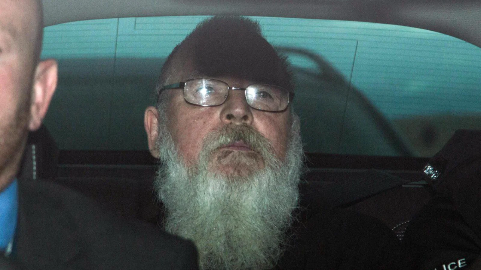 Angus Sinclair, white-bearded and wearing glasses, in the back of a police car