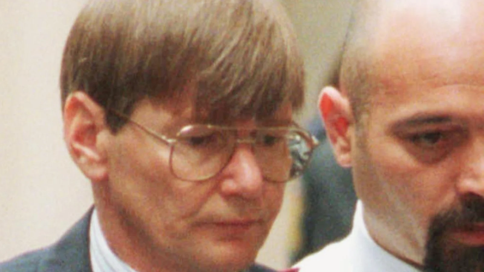 Peter Dupas in glasses and a suit, head lowered, beside a prison officer in a Melbourne court