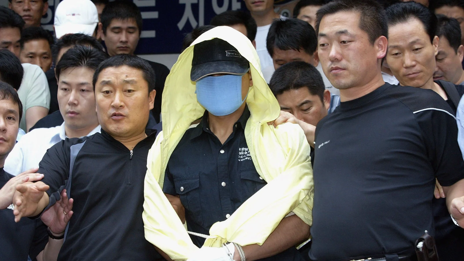 Yoo Young-chul, masked and hooded, led by plainclothes police officers through a crowd in Seoul