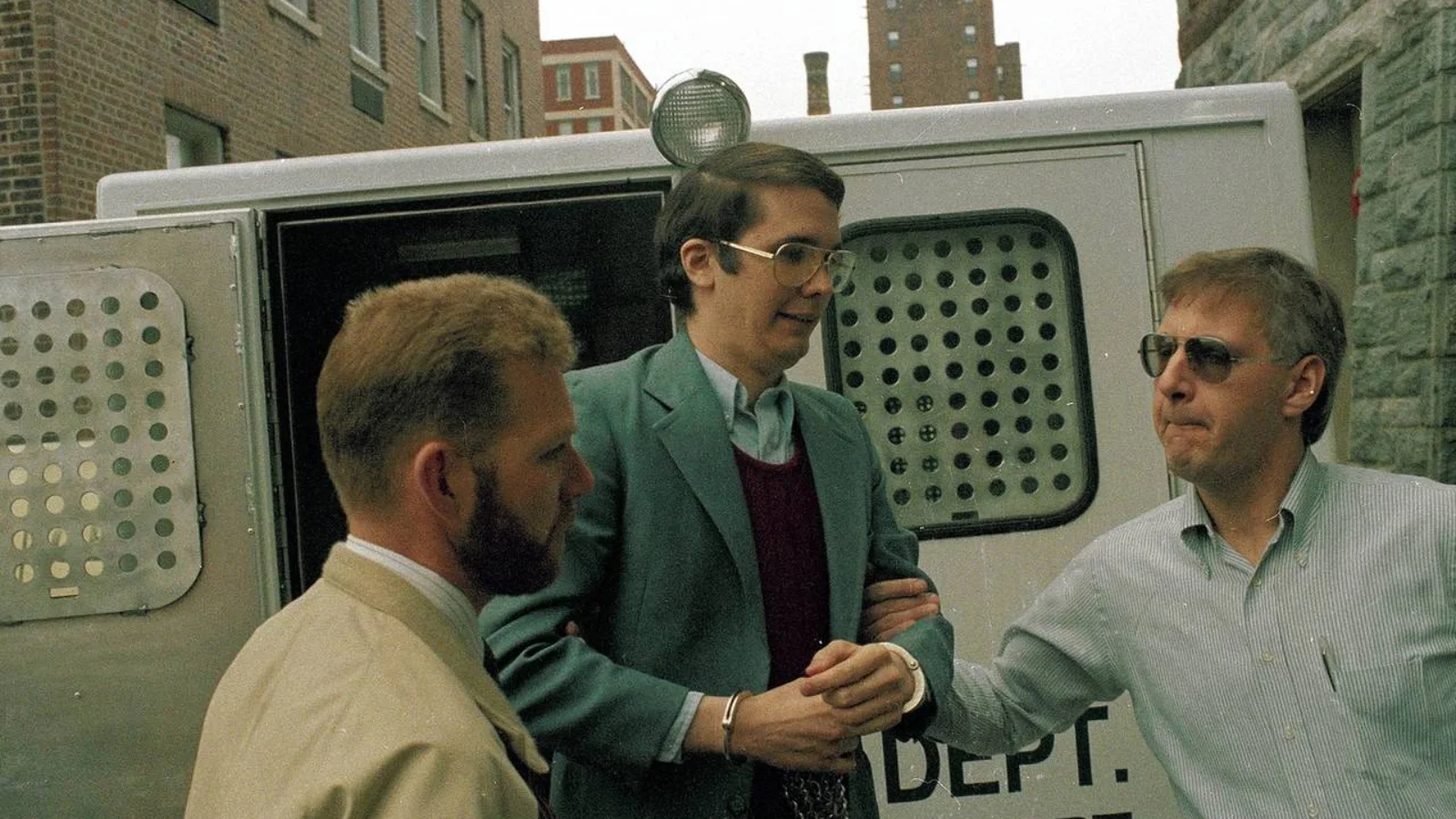 Michael Ross in glasses and a green jacket, handcuffed, led from a corrections transport van by two officers