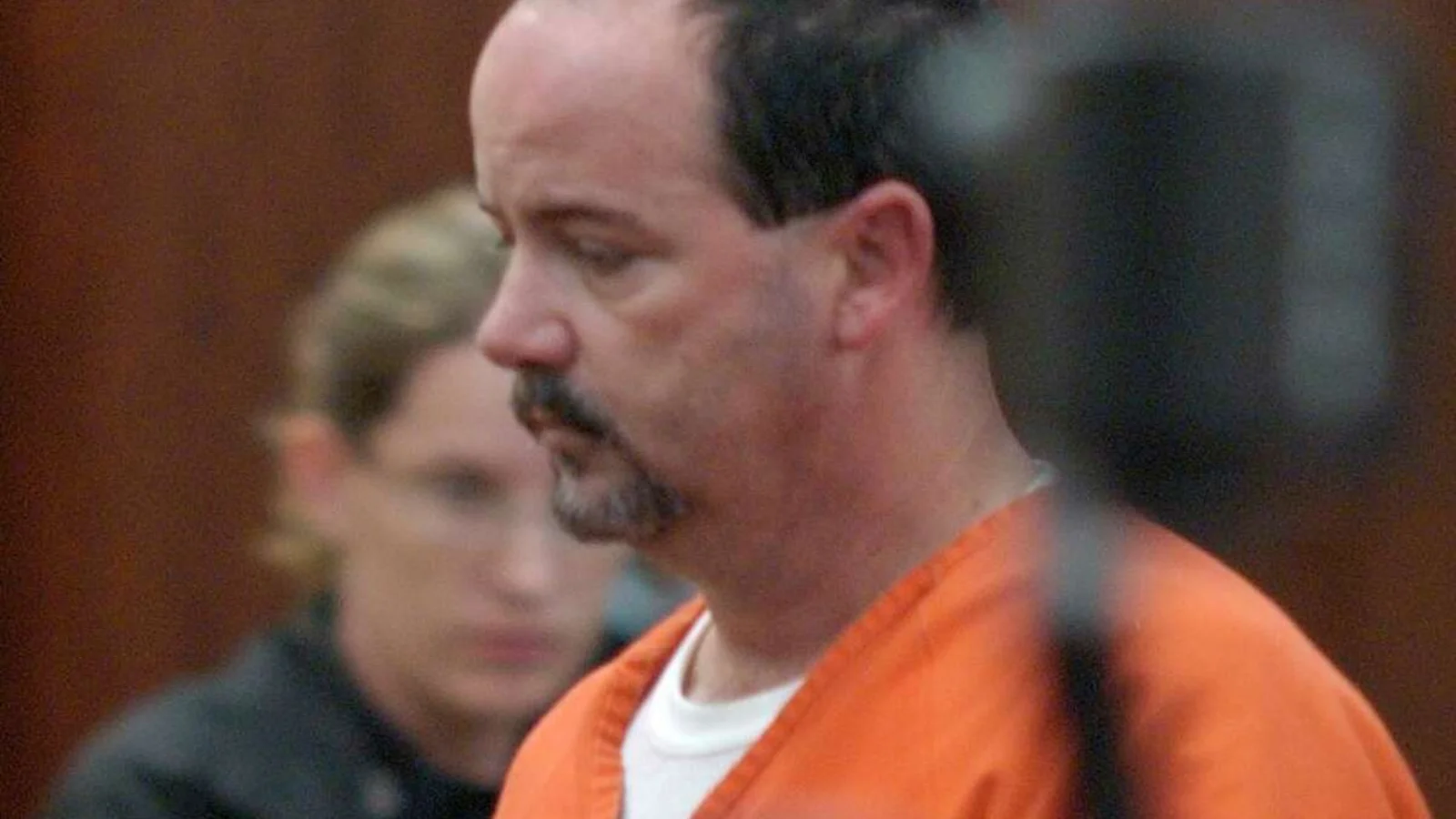 Anthony Shore in an orange jail jumpsuit standing in a Houston courtroom, a deputy behind him