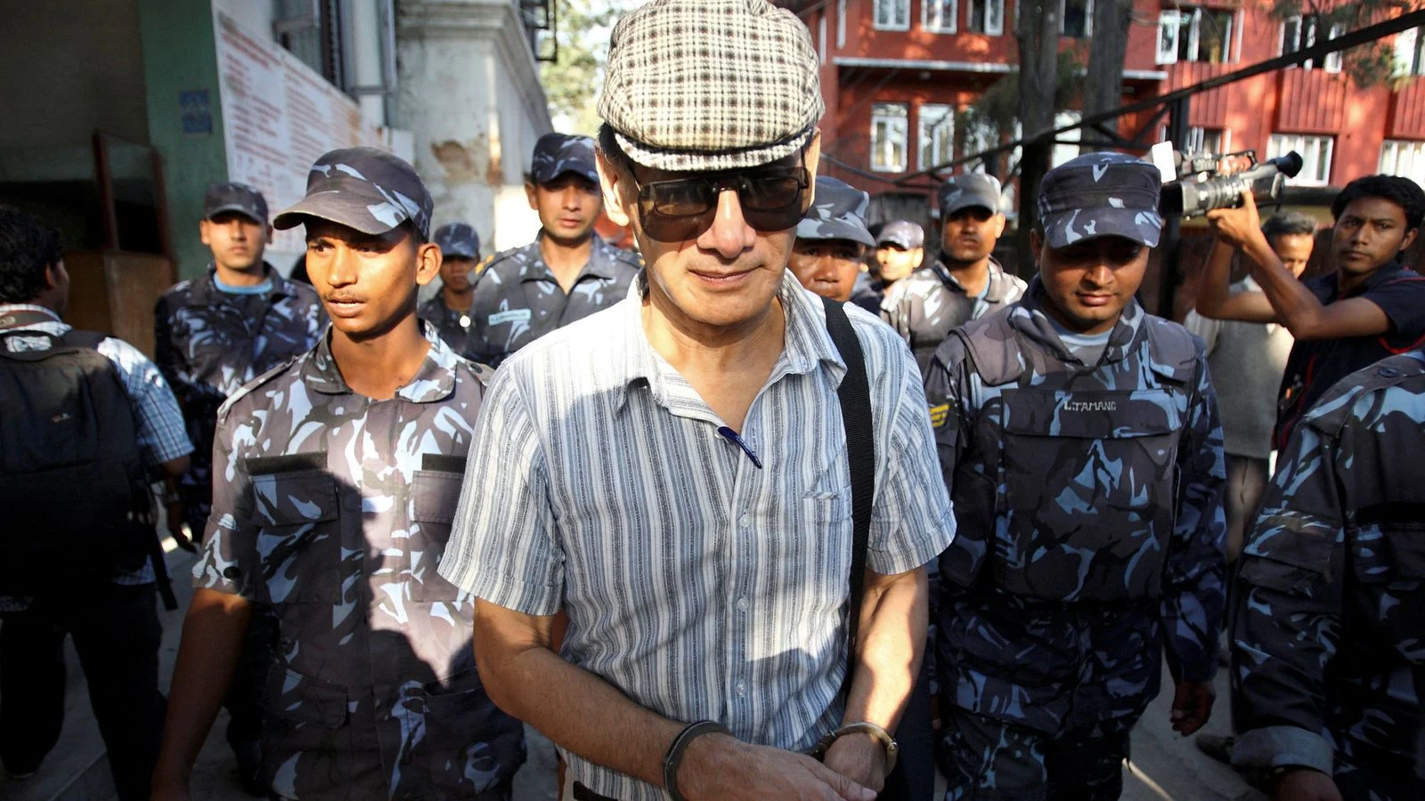 Charles Sobhraj in a flat cap and sunglasses escorted by armed Nepali police through a crowd in Kathmandu