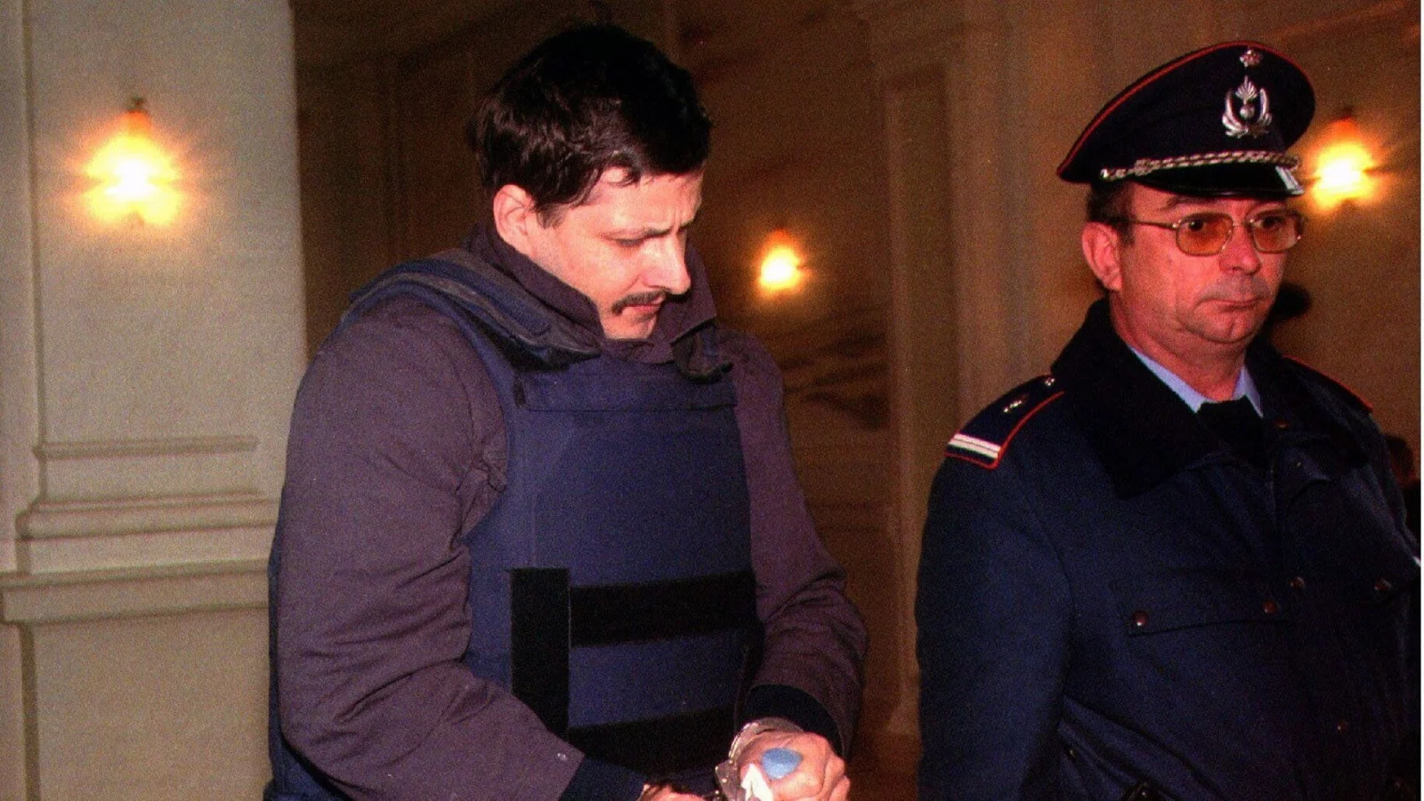 Marc Dutroux in a bulletproof vest, handcuffed, beside a uniformed Belgian police officer