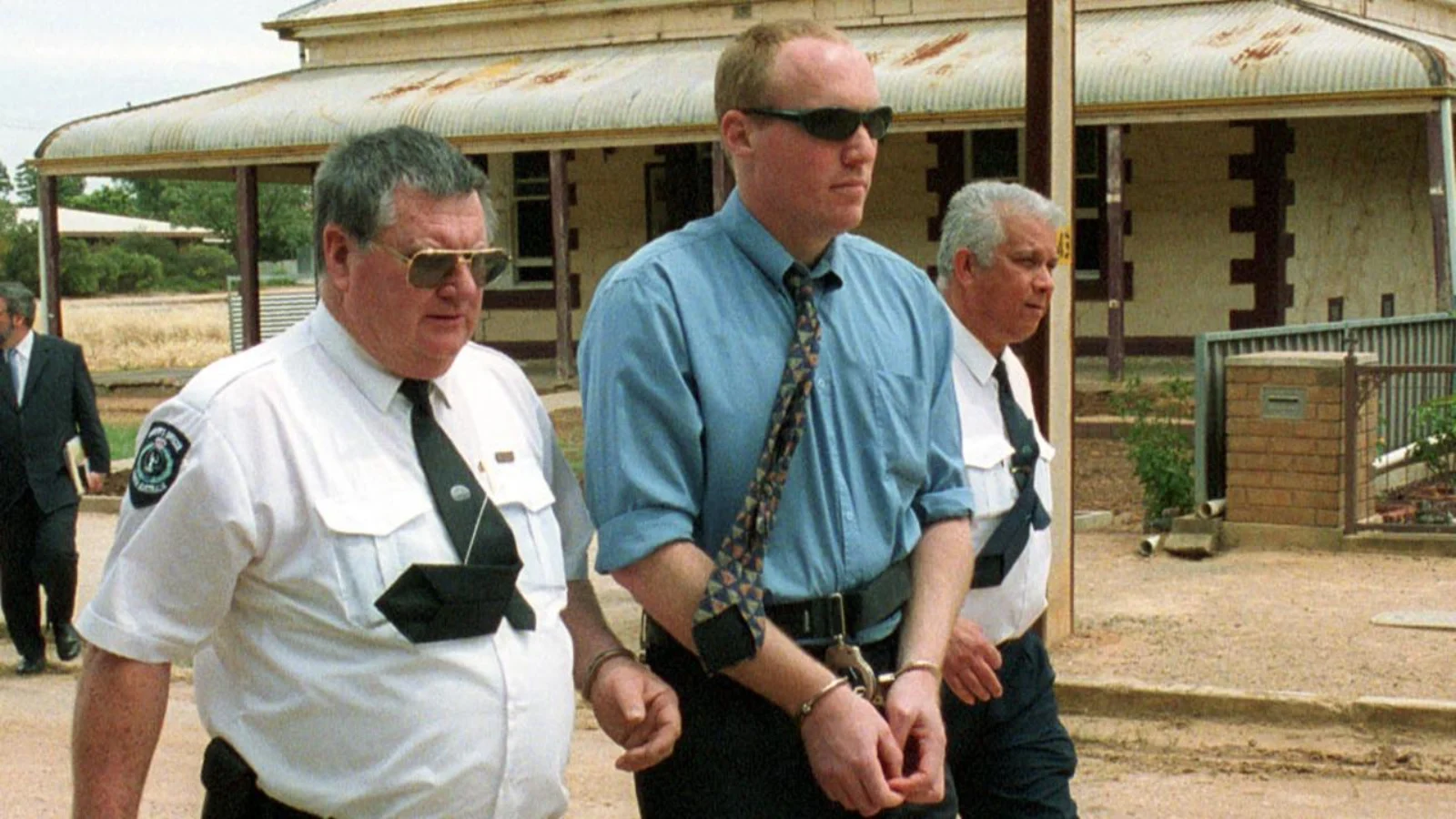 Robert Wagner in sunglasses and a blue shirt, handcuffed, escorted by South Australian police officers in Snowtown