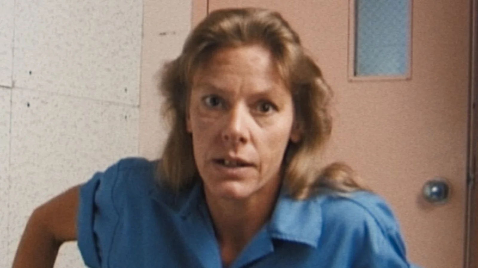 Aileen Wuornos in a blue prison shirt, leaning forward in a room with a pink door
