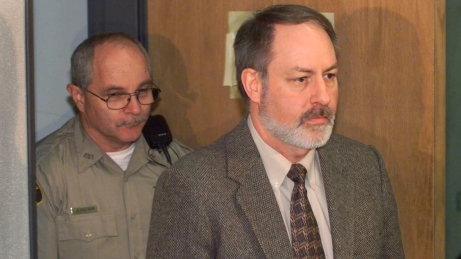 Robert Lee Yates Jr., gray-bearded, in a tweed jacket and tie, entering a courtroom ahead of a deputy