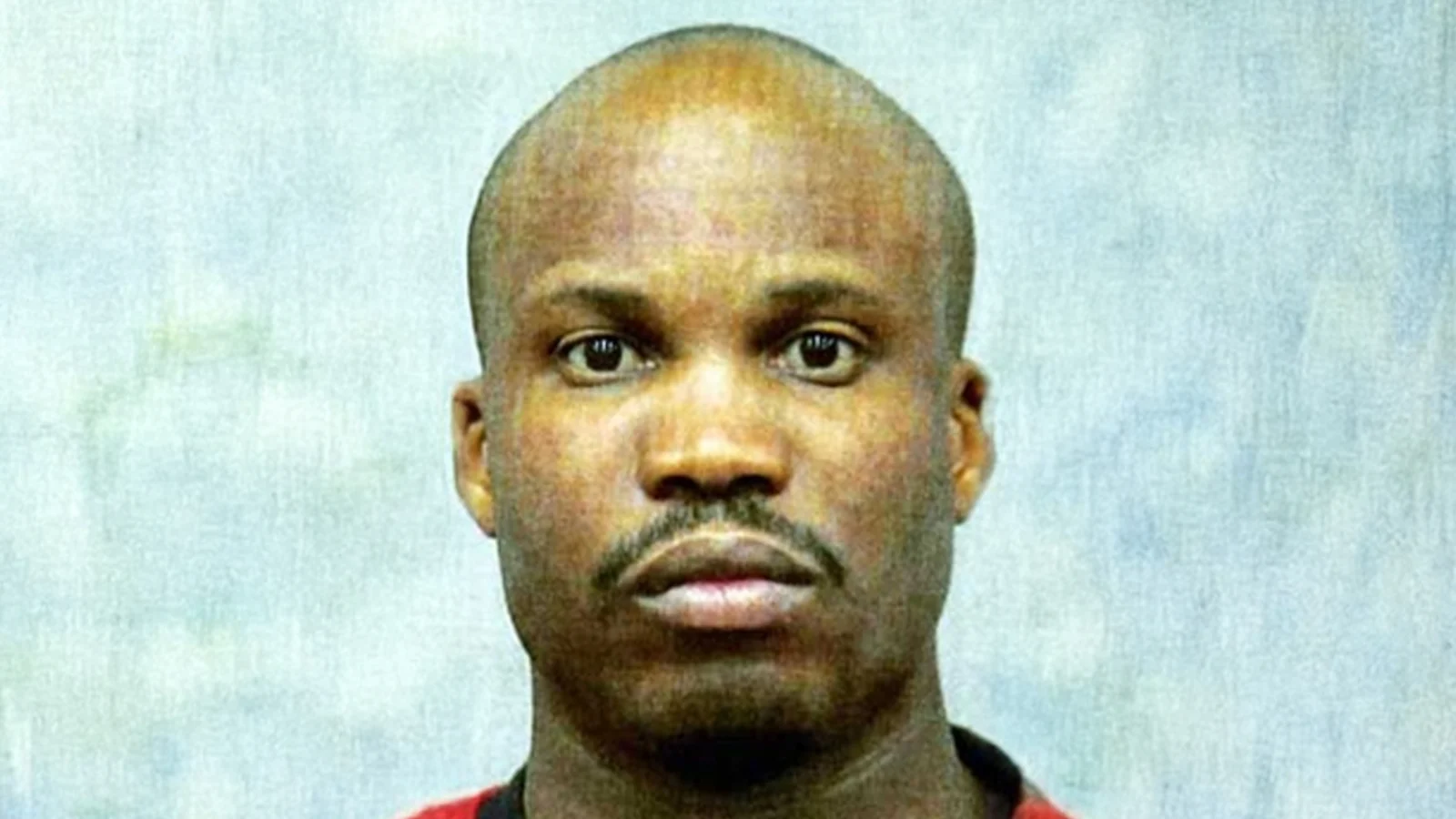 Booking photograph of Maury Travis, head shaved, in a red shirt against a pale blue backdrop