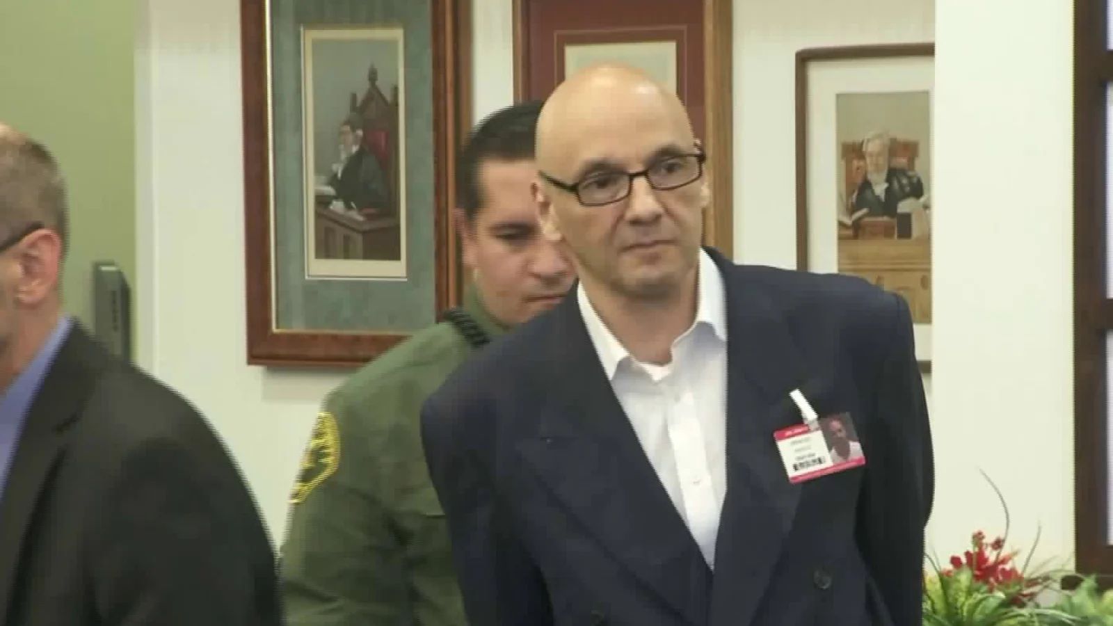 Andrew Urdiales, bald and in glasses, wearing a dark jacket and white shirt, led into a courtroom by a deputy