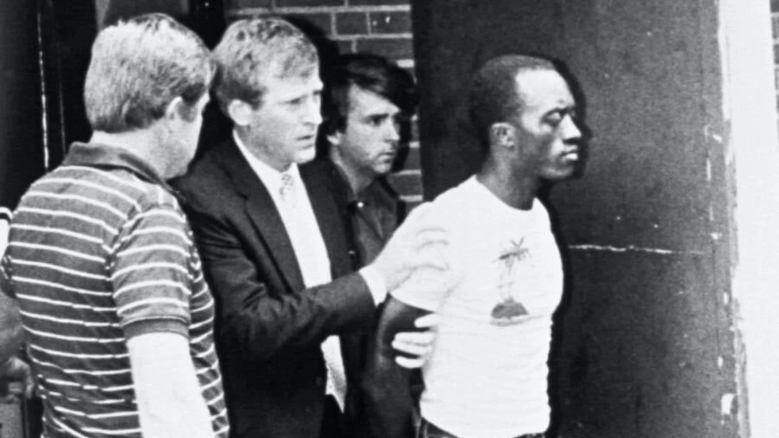 Alton Coleman, in a white T-shirt, held by officers outside a brick building after his 1984 arrest, in a black-and-white photograph