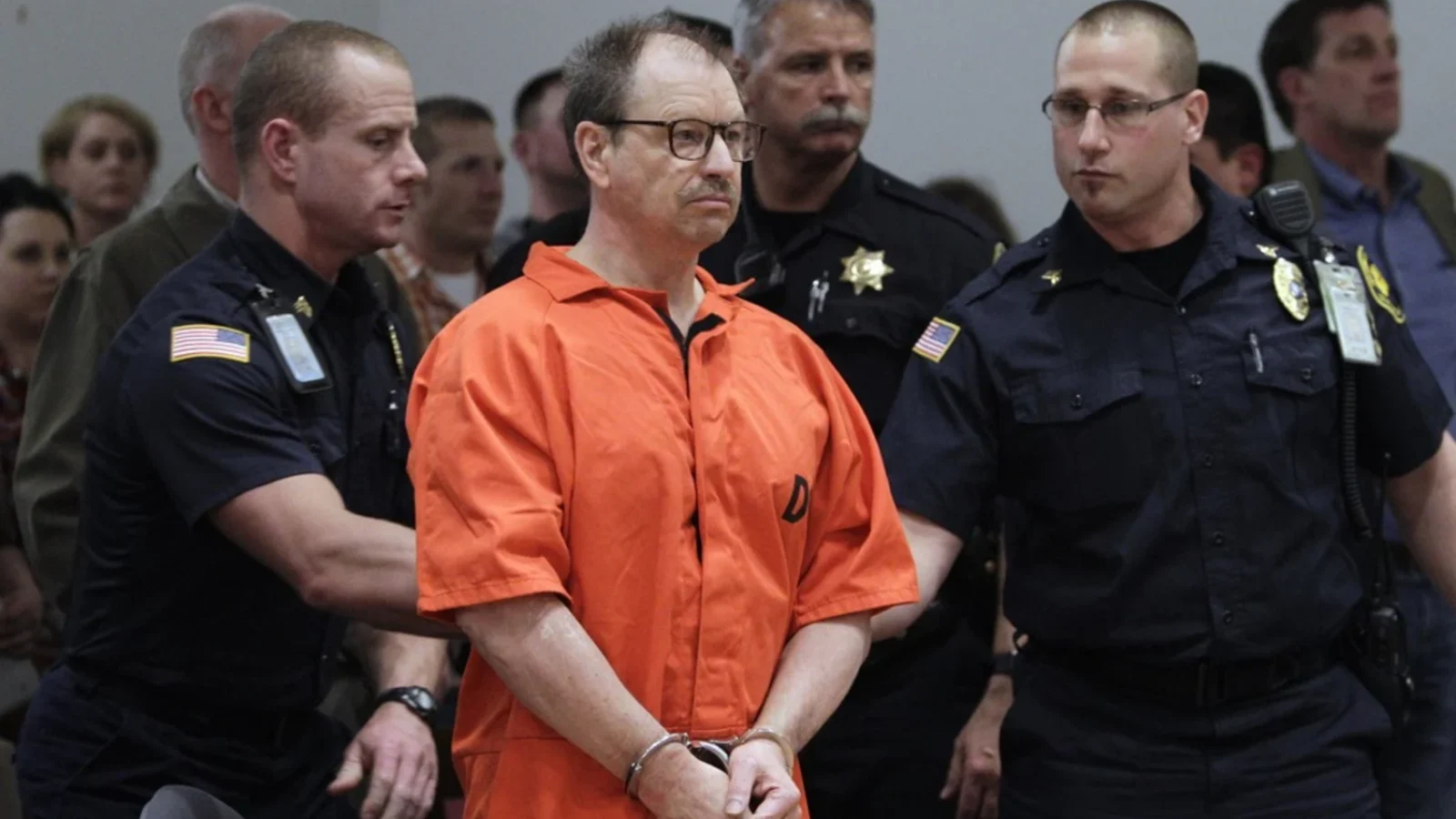 Gary Ridgway in an orange jail jumpsuit and glasses, handcuffed, standing in a King County courtroom with deputies behind him