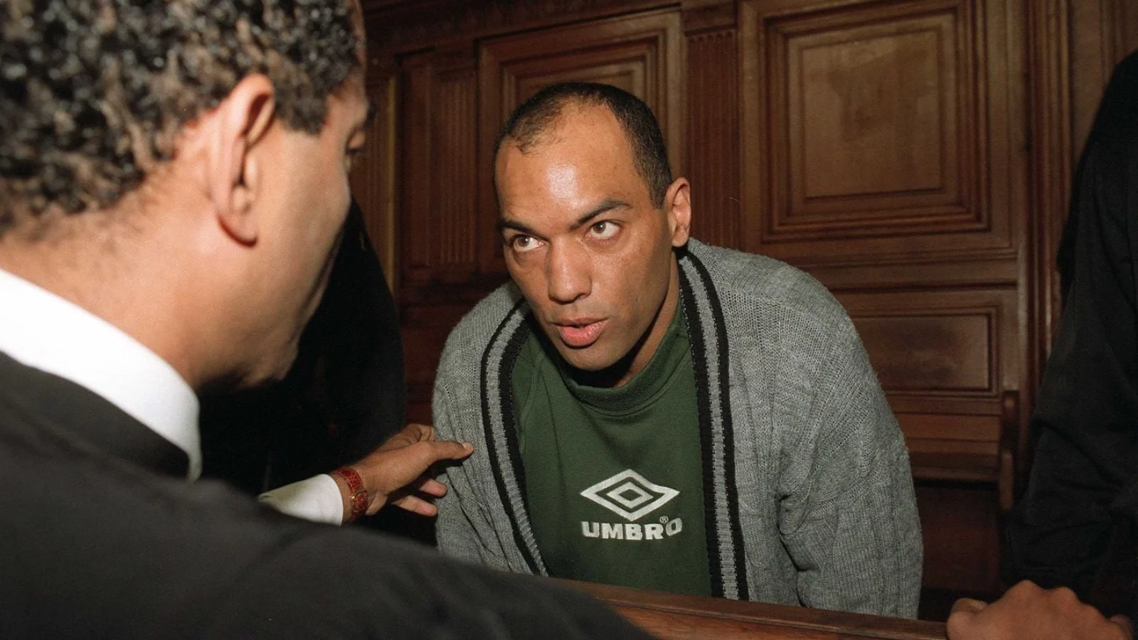 Guy Georges, head shaved, in a green sweater and gray cardigan, speaking with his lawyer in the dock of a Paris courtroom