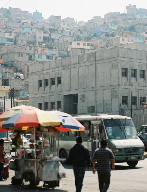 Illustration: a plain concrete public building in a hillside suburb, a street vendor's cart with umbrellas and a minibus in front, passers-by seen from behind. Not the actual courthouse.
