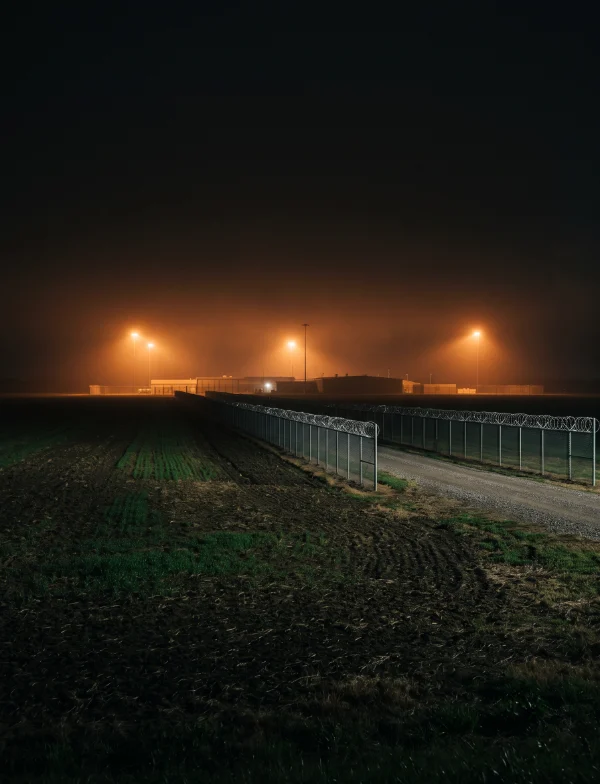 Illustration of a prison exterior under floodlights at night, a high fence in the foreground. Not the actual prison.