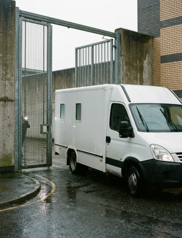 A white prisoner transport van at an open steel gate in a high concrete wall, on a wet road in the rain