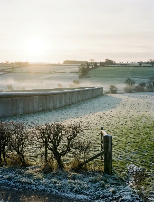A high curved-top perimeter wall crossing frosted green fields at sunrise, with a bare hedge and a gatepost in the foreground. Not the actual prison.