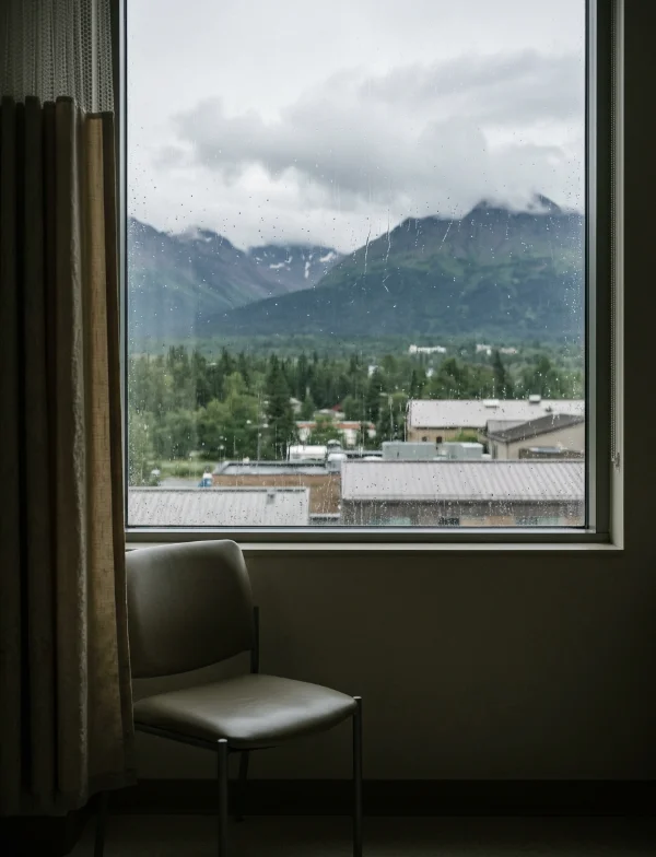 Illustration of a rain-streaked hospital window looking out on spruce trees, low roofs and clouded Alaska mountains, with an empty chair. Not the actual hospital.