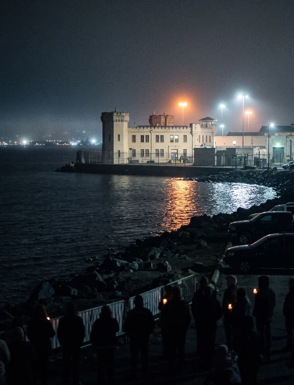Illustration of protesters holding candles on a bayside path facing a lit prison building across the water at night. Not an actual scene from the case.