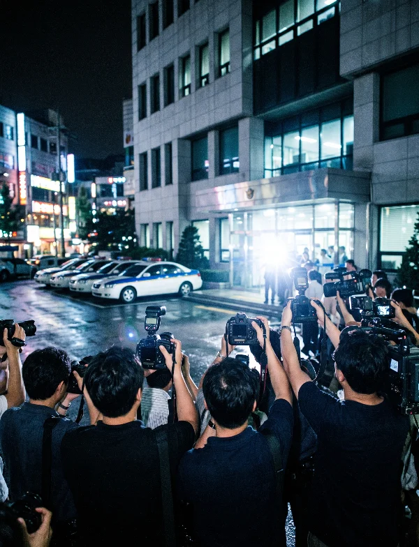 Illustration of press photographers crowding outside a Seoul police station at night under camera flashes. Not an actual scene from the case.